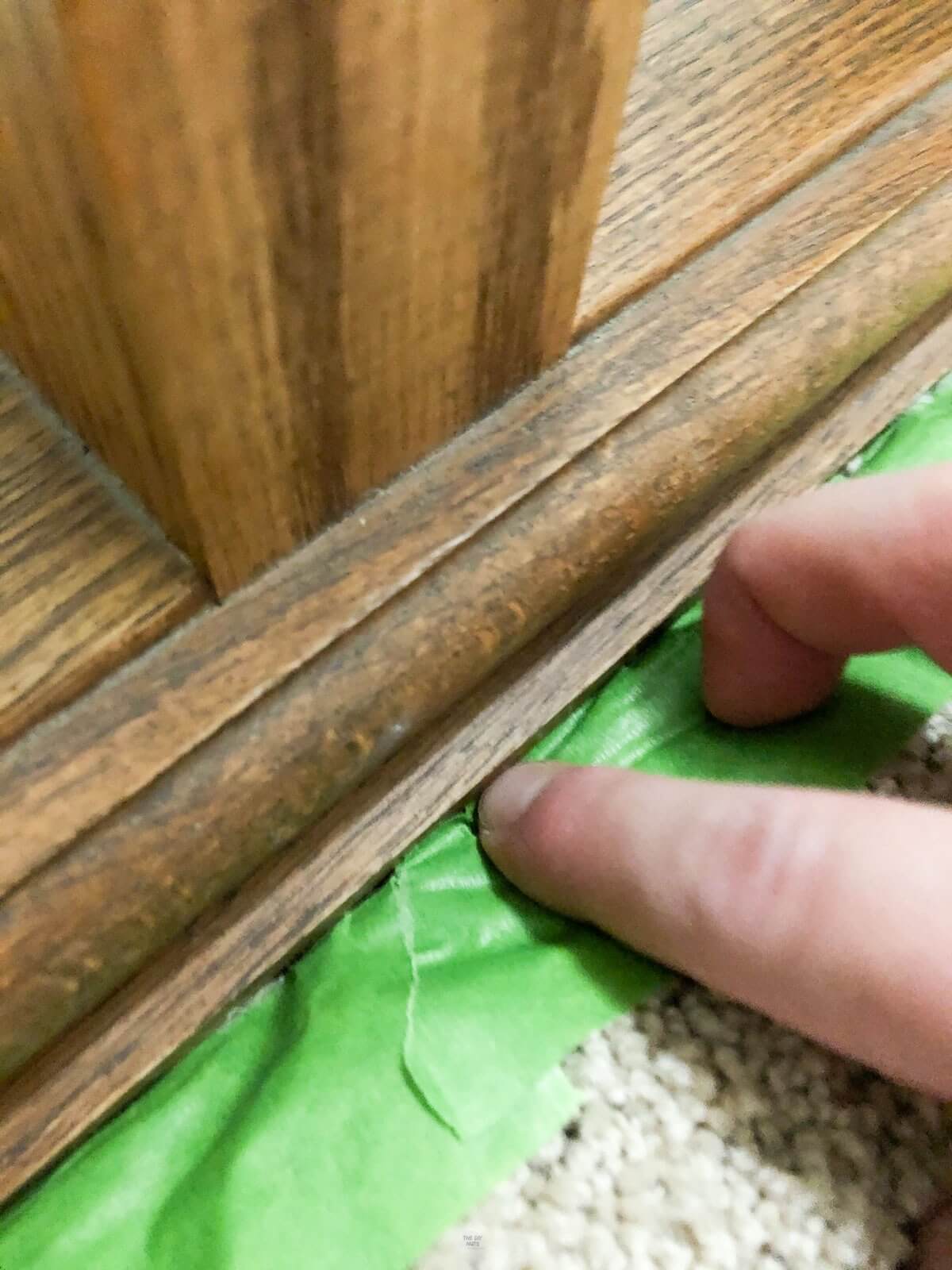 fingers pushing down green painter's tape on carpet under oak stair railing.