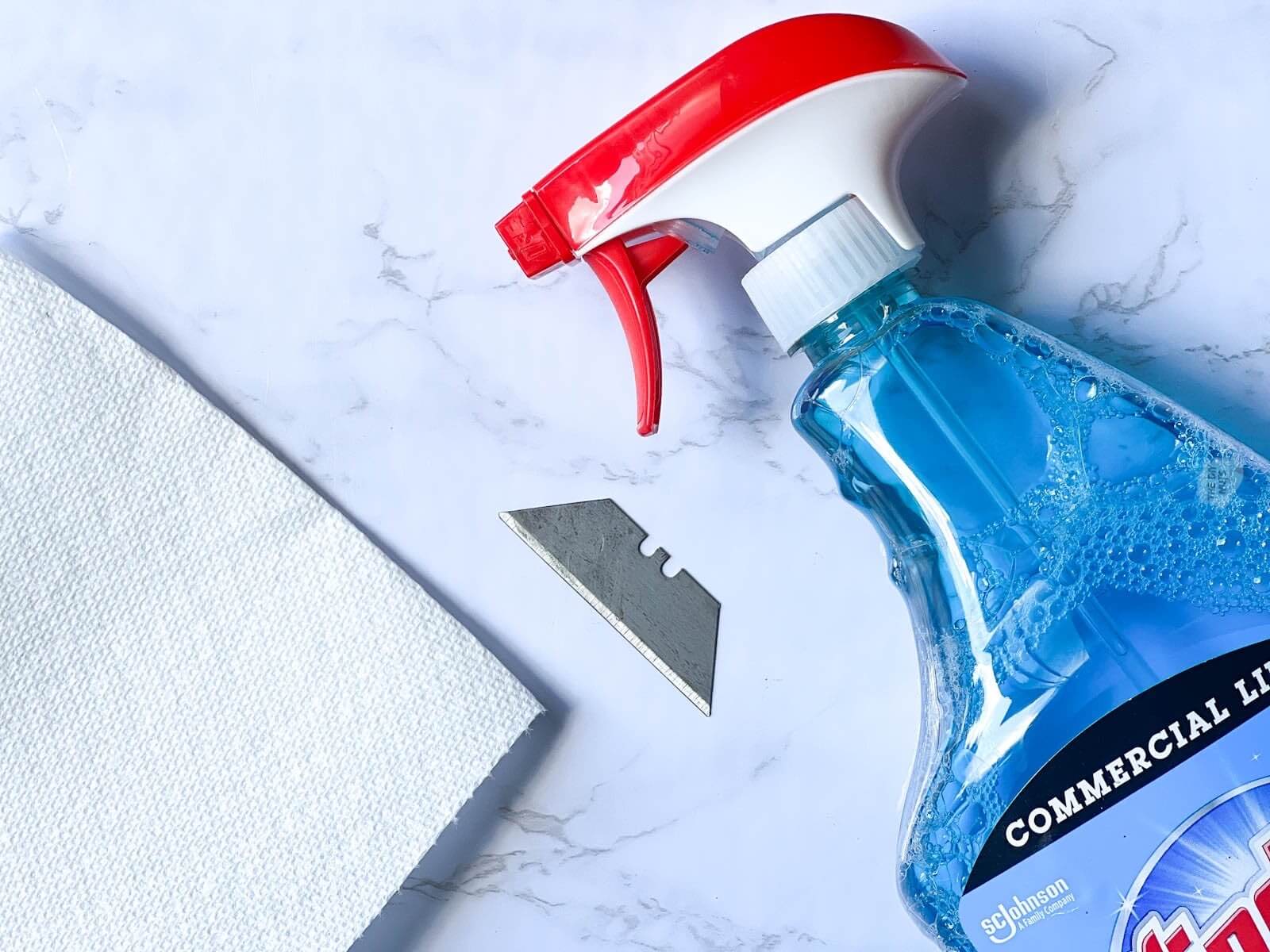 glass cleaner in spray bottle, razor blade and paper towel on marble countertop.