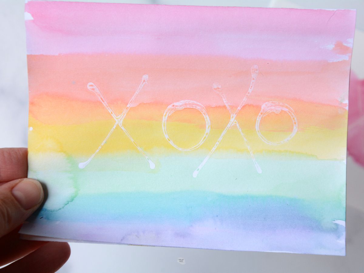hand holding rainbow watercolor card with xoxo written in glue.