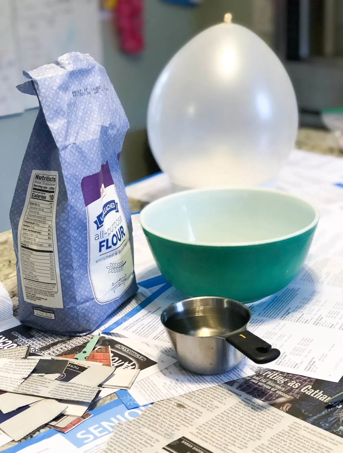 flour, balloon, bowl, measuring cup filled with water on newspaper in kitchen.