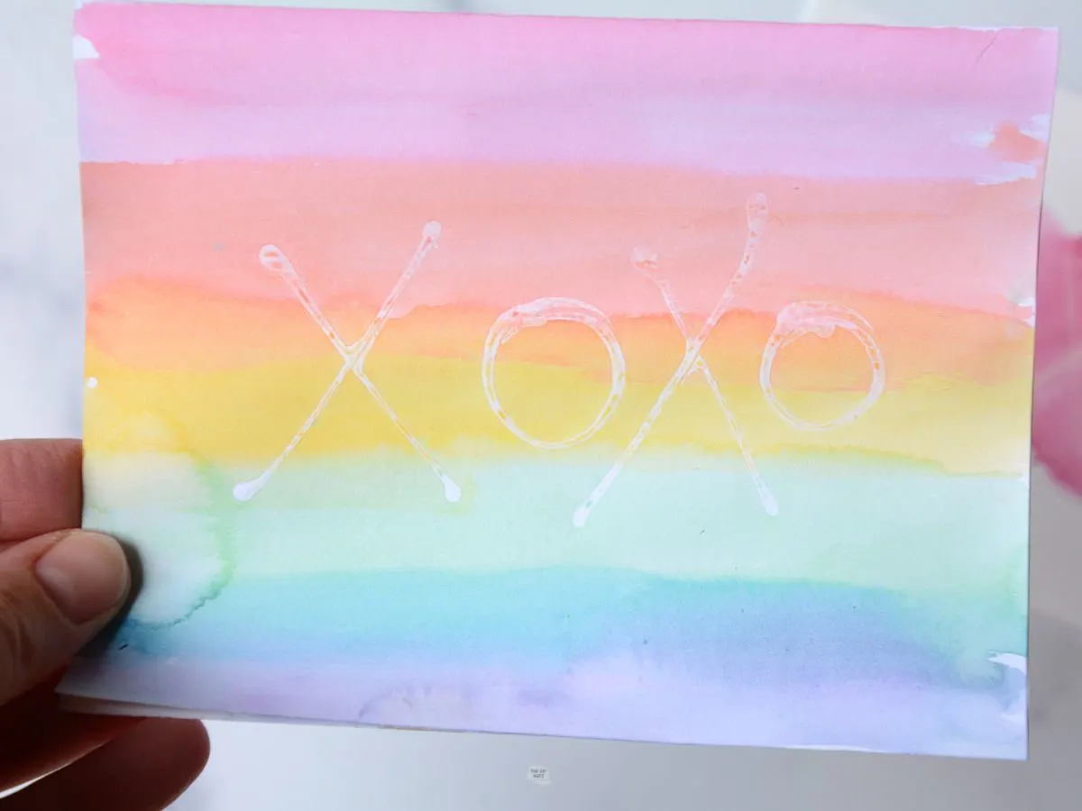 hand holding rainbow watercolor card with xoxo written in glue.